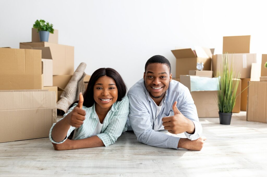 Happy black couple recommending real estate agency, showing thumbs up while lying on floor among