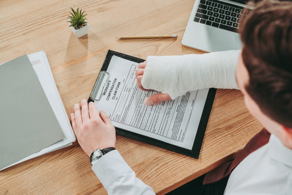 cropped view of worker holding broken arm compensation claim and sitting at table in office,